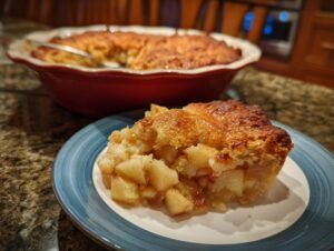 A close-up view of a slice of warm apple pie dump cake on a blue-rimmed plate, with the rest of the cake in a red baking dish in the background.