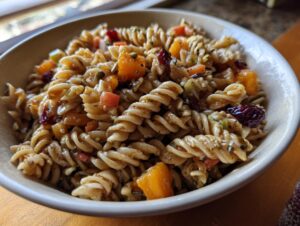 A close-up of a bowl filled with a hearty fall pasta salad, featuring fusilli pasta, butternut squash cubes, dried cranberries, and herbs.