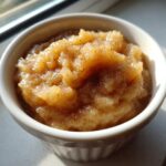 A close-up of a white ramekin filled with chunky homemade applesauce, dusted with cinnamon.
