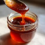 Close-up of thick, glossy honey BBQ sauce dripping from a spoon into a glass jar.