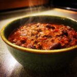 A close-up shot of a steaming bowl of hearty chili, featuring ground beef and kidney beans in a rich tomato sauce.