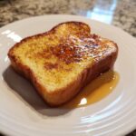 A single slice of golden-brown protein french toast drizzled with syrup on a white plate.