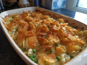 A close-up of a freshly baked Tuna Casserole in a white baking dish, topped with crushed potato chips and peas.