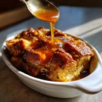 A spoonful of rich custard sauce being drizzled over a serving of golden-brown bread pudding in a white dish.