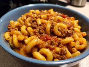 A close-up of a blue bowl filled with delicious American goulash, featuring elbow macaroni and seasoned ground beef in a rich tomato sauce.