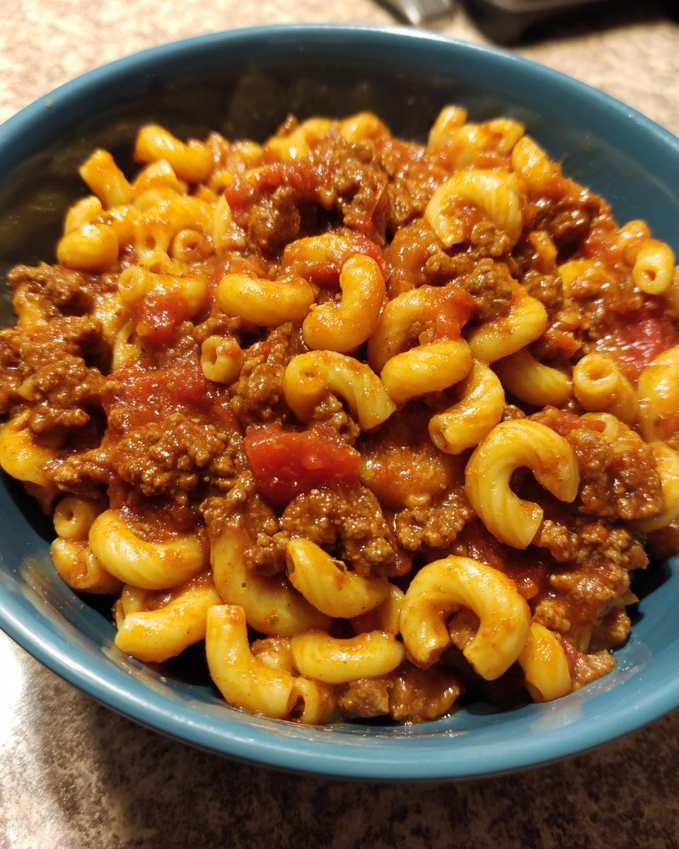 A close-up of a blue bowl filled with American goulash, featuring elbow macaroni and seasoned ground beef in a rich tomato sauce.
