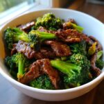 Close-up of a white bowl filled with tender slices of beef and vibrant green broccoli florets coated in a glossy sauce.