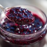 A close-up of a spoon filled with glistening homemade blueberry jam, held over a jar of the same jam.