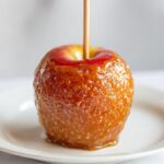A close-up of a shiny, golden-brown caramel apple on a white plate, ready to be enjoyed.