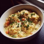 A close-up of a white bowl filled with creamy chicken and dumplings, garnished with fresh parsley.