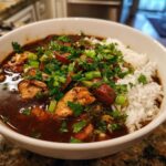 A close-up of a white bowl filled with delicious chicken and sausage gumbo served over rice, garnished with fresh parsley and green onions.