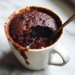A close-up of a warm chocolate mug cake with a gooey molten center, a spoon taking a bite.