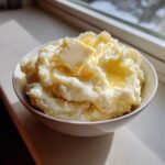 A bowl of fluffy mashed potatoes topped with a melting pat of butter and a drizzle of butter.
