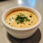 A close-up of a white bowl filled with creamy queso dip, garnished with fresh chopped cilantro and red pepper flakes.