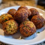 Close-up of a pile of golden-brown, crispy falafel balls with visible herbs on a white plate.