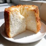 A close-up of a fluffy Angel Food Cake slice on a white plate, with a golden-brown crust.