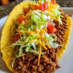 A close-up of a delicious ground beef taco filled with seasoned ground beef, shredded lettuce, diced tomatoes, and shredded cheese.