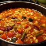 A close-up of a pot filled with hearty Minestrone soup, featuring pasta, beans, and colorful vegetables.