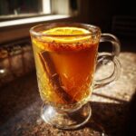 A close-up of a glass mug filled with warm mulled apple cider, garnished with an orange slice, cinnamon stick, and spices.