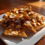 Close-up of crunchy peanut brittle pieces scattered on a white plate, showcasing peanuts in a golden caramel.