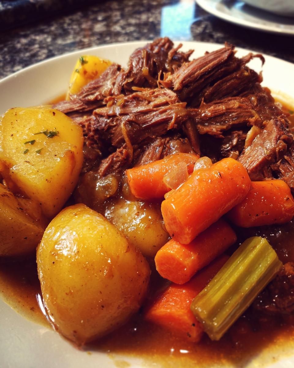 A close-up of a comforting plate of pot roast, tender shredded beef, golden potatoes, and bright orange carrots in savory gravy.