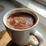 Close-up of a steaming mug of rich hot chocolate with frothy top, bathed in natural light.