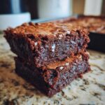 A close-up of two stacked, fudgy brownies topped with flaky sea salt, showcasing their rich chocolate texture.