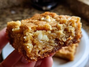 A close-up shot of a chewy blondie bar studded with white chocolate chips, held in a hand.