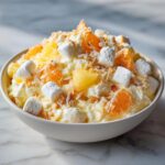 A close-up of a bowl filled with fluffy ambrosia salad, featuring marshmallows, mandarin orange segments, pineapple chunks, and shredded coconut.