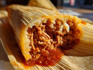 Close-up of a freshly made tamale, unwrapped to reveal tender shredded pork in red sauce and fluffy masa.