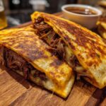 Close-up of two golden-brown quesadillas filled with shredded birria meat and melted cheese, served with consommé.