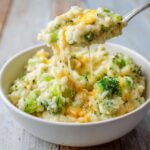 A spoonful of creamy broccoli rice being lifted from a white bowl, showing melted cheese strings.