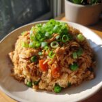 A close-up of a bowl of chicken fried rice, topped with fresh green onions, peas, and carrots.