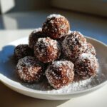 A mound of rich, dark chocolate rum balls generously coated in white powdered sugar on a white plate.