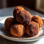 A pile of rich chocolate truffles generously dusted with cocoa powder on a white plate.