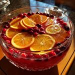 A close-up of a festive Christmas punch in a glass bowl, garnished with bright orange slices and fresh cranberries.