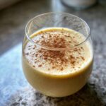A close-up of a glass filled with creamy eggnog, topped with a sprinkle of cinnamon.