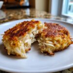 Close-up of two golden-brown, perfectly cooked crab cakes on a white plate, one split open to reveal flaky crab meat.