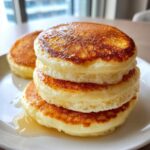 A close-up of a stack of three fluffy pancakes drizzled with syrup on a white plate.