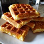 A close-up stack of freshly made, golden brown Belgian waffles resting on a white plate.