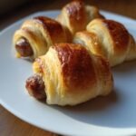 Close-up of four freshly baked, golden brown Pigs in a blanket served on a white plate.
