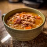 A close-up of a steaming bowl of Ham and bean soup filled with white beans, shredded ham, and diced carrots.