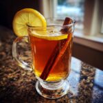 A glass mug filled with a warm Hot Toddy, garnished with a lemon slice and a cinnamon stick.