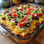 A close-up of a freshly made Seven Layer Dip in a glass baking dish, topped with tomatoes, olives, and green onions.