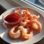 A plate of chilled, cooked shrimp arranged around a small glass bowl of bright red cocktail sauce, ready for serving as shrimp cocktail.