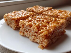 Close-up of several golden brown, gooey Rice Krispie treats cut into squares and resting on a white plate.