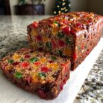 A moist holiday fruitcake with a slice cut, showcasing colorful candied fruits and nuts.