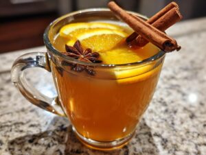 Close-up of a glass mug filled with warm spiced apple cider, garnished with orange slices, cinnamon sticks, and star anise.