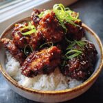 A bowl of fluffy white rice topped with glistening, glazed Teriyaki chicken pieces, garnished with sesame seeds and green onions.