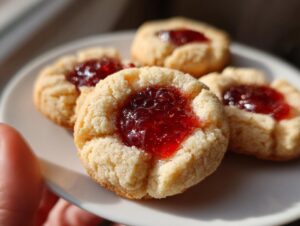 Close-up of four classic thumbprint cookies filled with glistening red jam on a white plate.